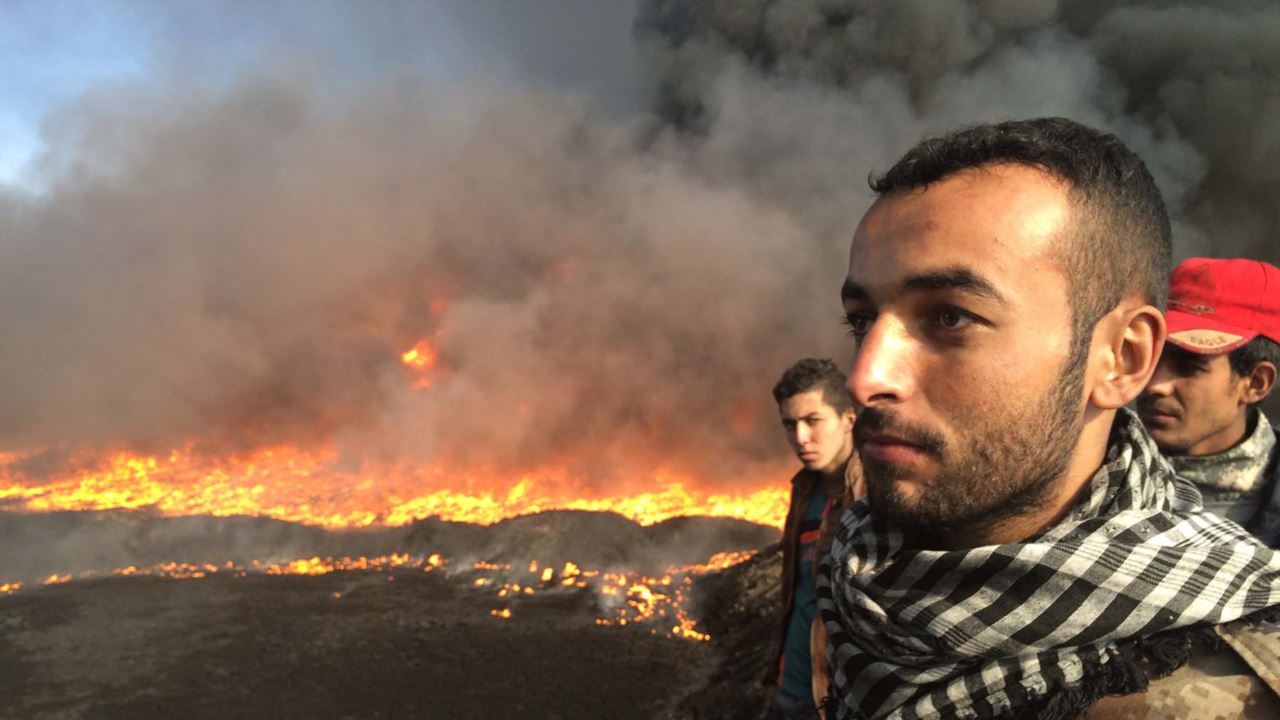Men standing against a horizon of burning oil fields.